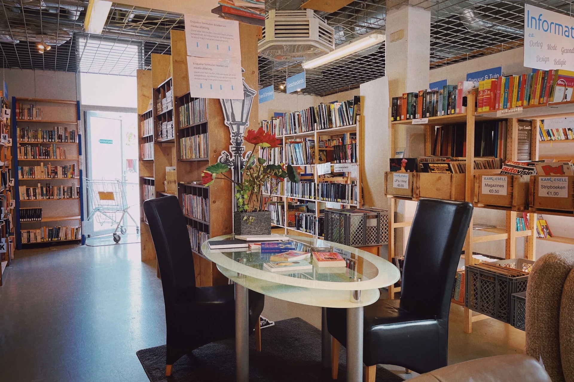 Binnenaanzicht kringloopwinkel met boekenrekken, leeshoek met tafel en stoelen, en plantenversiering