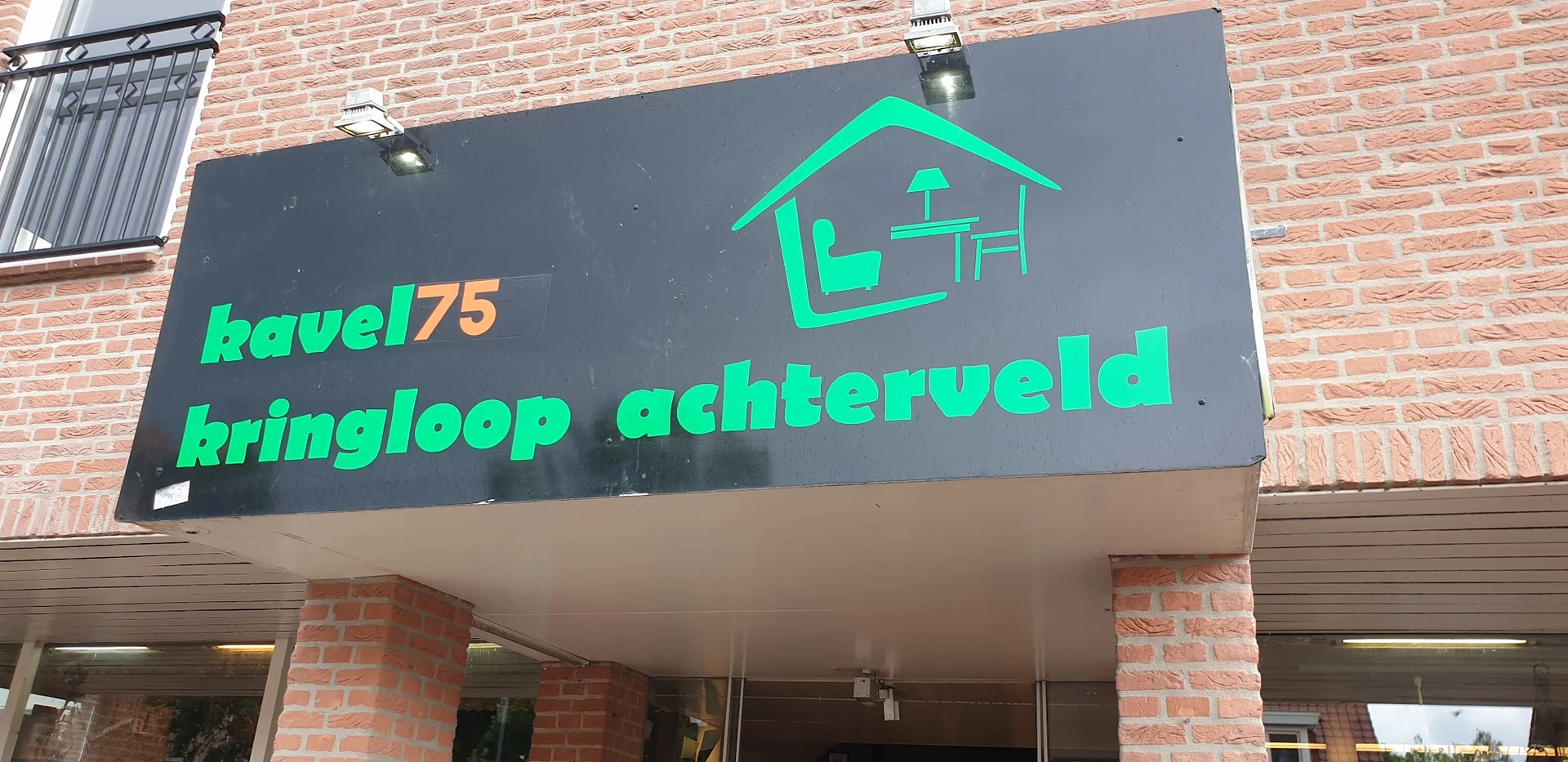 Grijze gevel met groen neonbord van kringloopwinkel met huisicon en tekst 'ingloop achterveld'