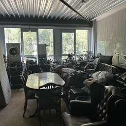 Interieur van kringloopwinkel met tafels, stoelen en diverse gebruikte spullen langs grote ramen
