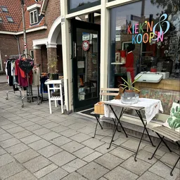 Voorkant van kringloopwinkel met witte stoelen en tafel op terras, kledingrek buiten, geopende deur en raam met kleurrijke le