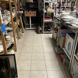 Binnenaanzicht van kringloopwinkel met tegelvloer, stellingen met serviesgoed aan beide zijden en diverse kastjes en apparatu