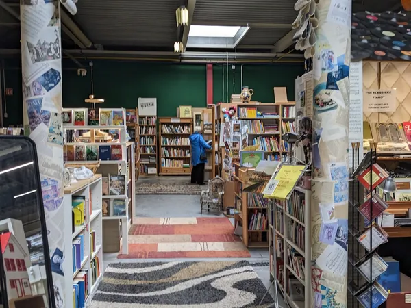 Binnenaanzicht van een kringloopwinkel met boekenkasten, stellingen en displays langs de zijkanten en een persoon die rondloo