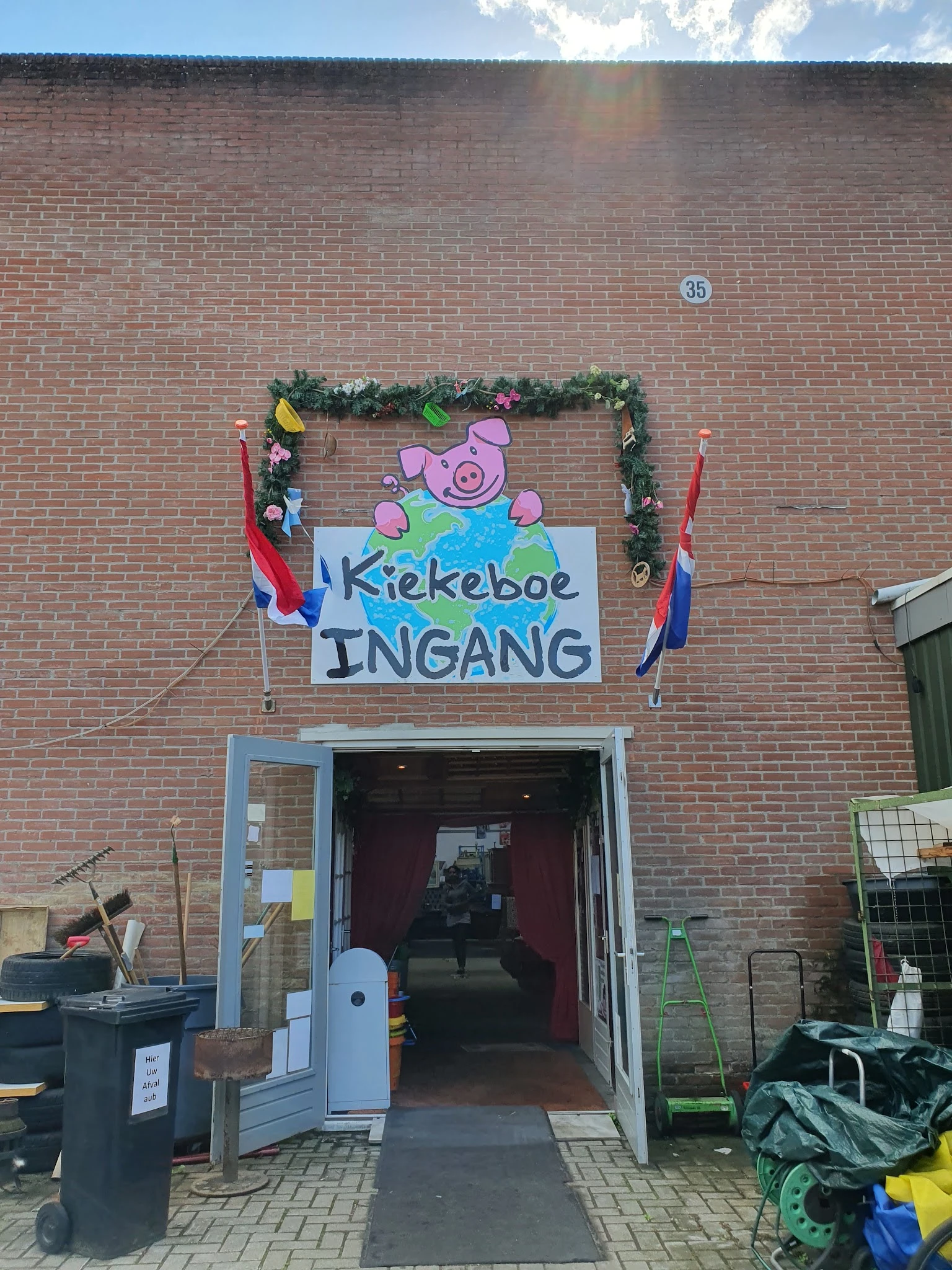 Bakstenen gevel met ingang en beschilderd welkomstbord met varken en Nederlandse vlag