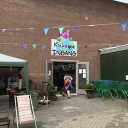 Gevel van kringloopwinkel Kiekeboe met naam op bord, versierd met bunting, ingang zichtbaar met persoon in deuropening