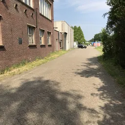 Zijkant van een bakstenen gebouw met daarnaast een betonnen inrit langs tuinhaag