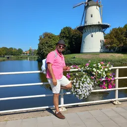 Man in roze shirt poseert aan railing met bloemen en molen op achtergrond