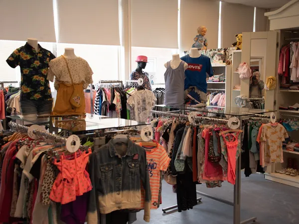 Binnenaanzicht van kledingwinkel met rekken en toonbanken vol tweedehands kleding, poppen met kledij en grote ramen.