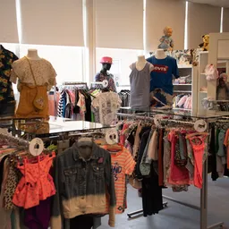 Binnenaanzicht van kledingwinkel met rekken en toonbanken vol tweedehands kleding, poppen met kledij en grote ramen.