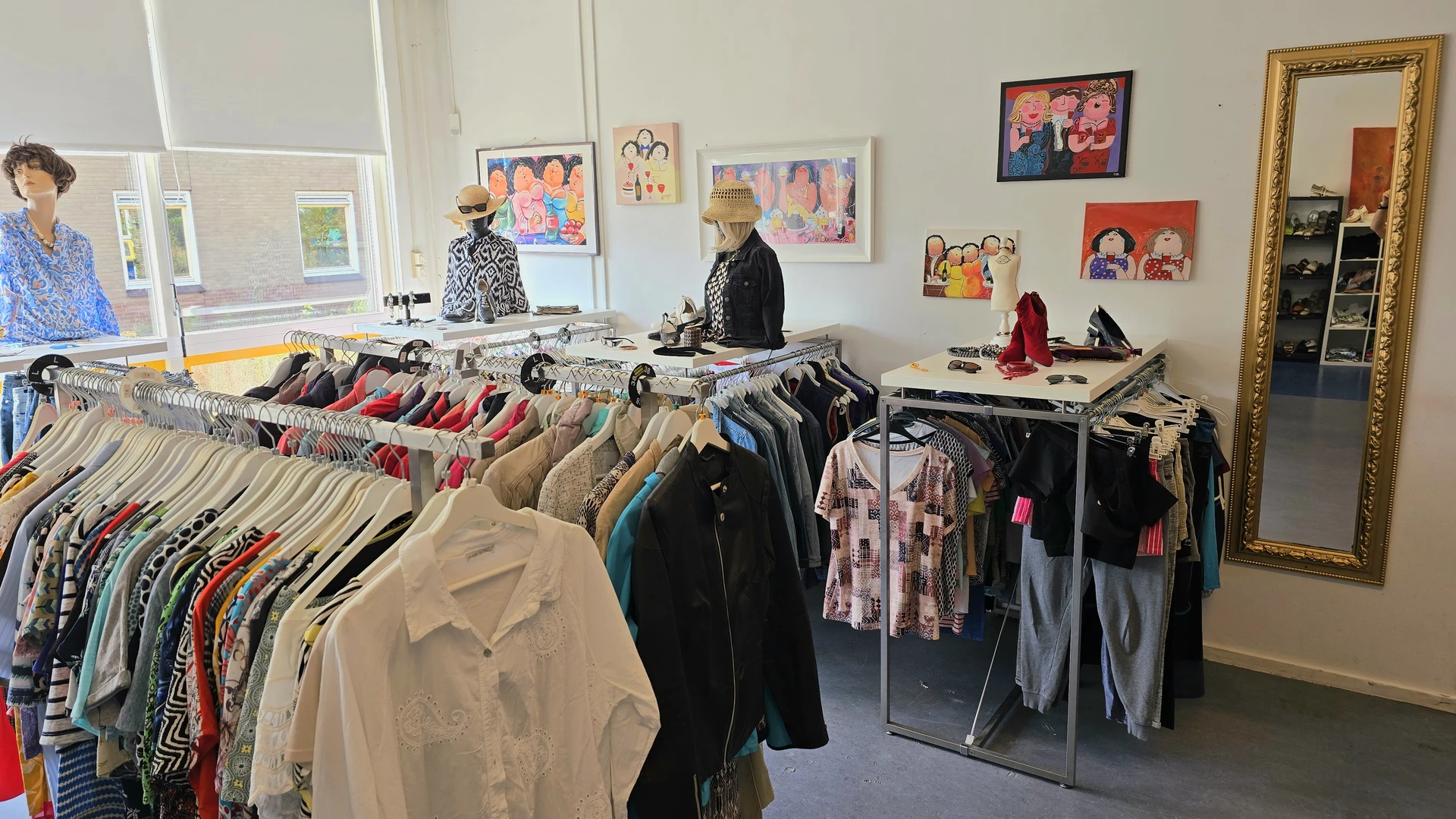 Winkelinterieur met kledingrekken vol tweedehands kleding, schilderijen aan de muur en mannequins met outfits