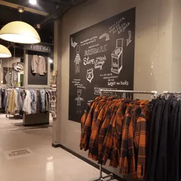 Winkelvloer met kledingrekken, hangende kledij in oranje en zwart, pendant lampen en decoratief schoolbord aan de muur