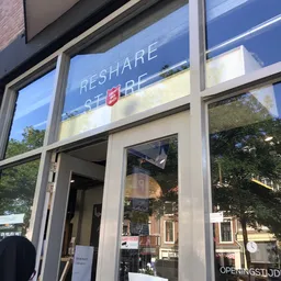 Glazen winkelpui met 'RESHARE' tekst en logo op raam, ingang zichtbaar met groene beplanting ernaast