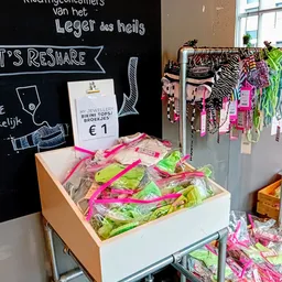 Uitgestalde tweedehands kleding in felle kleuren op tafels en aan rekken, met een bordje 'Let's Reshape' en prijskaartje van 