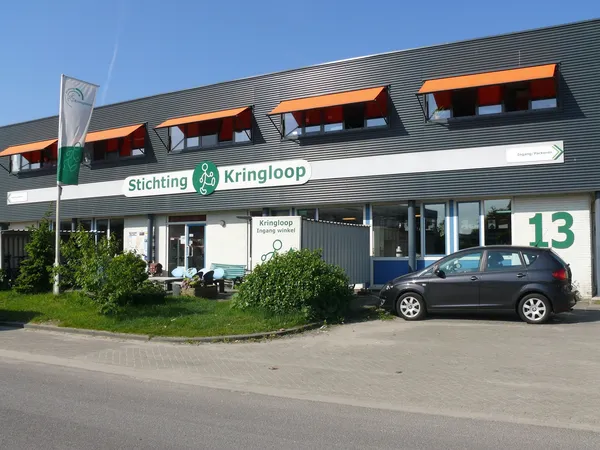 Gevel van Stichting Kringloop kledingwinkel met oranje dakramen en nummer 13, parkeerplaats met auto ervoor