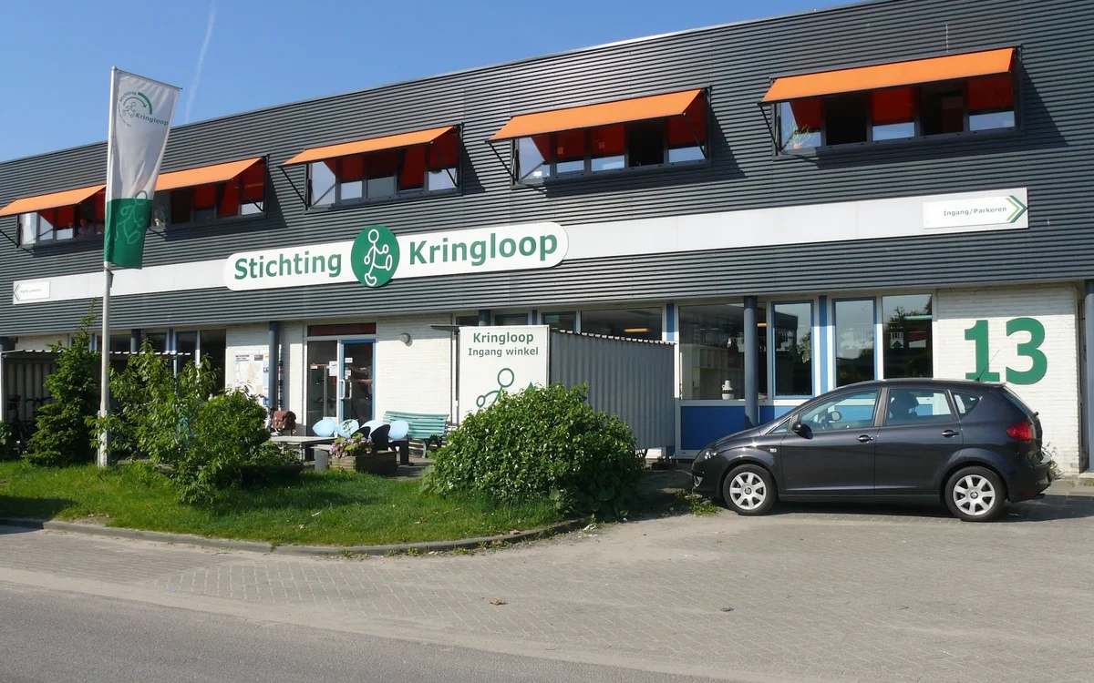 Gevel van Stichting Kringloop kledingwinkel met oranje dakramen en nummer 13, parkeerplaats met auto ervoor