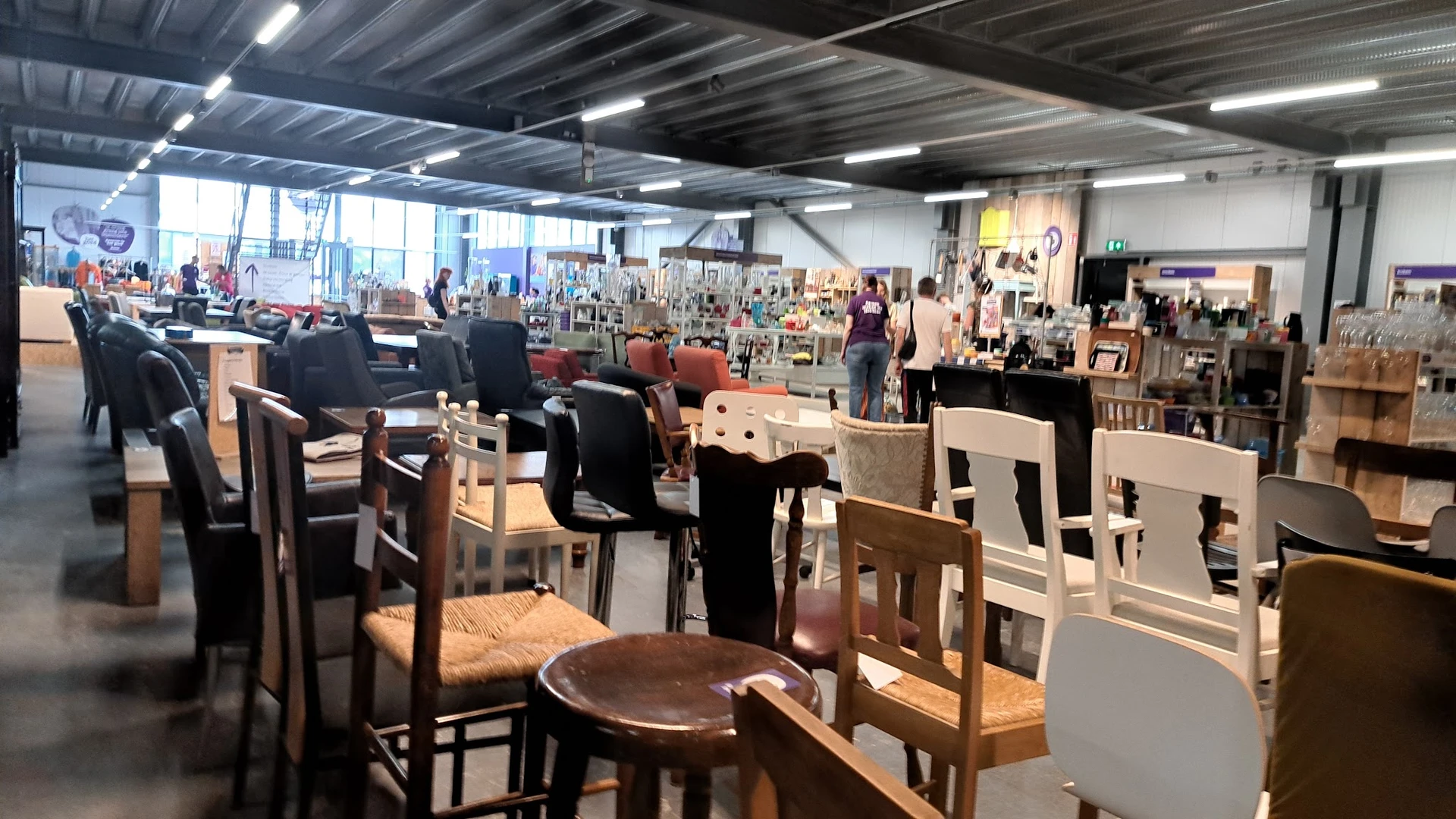 Ruim interieur van kringloopwinkel met verschillende meubels, stoelen en tafels uitgestald, fluorescente verlichting, veel be