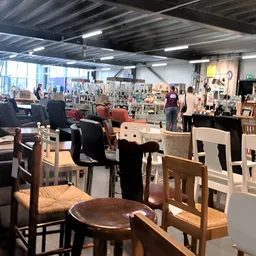 Ruim interieur van kringloopwinkel met verschillende meubels, stoelen en tafels uitgestald, fluorescente verlichting, veel be