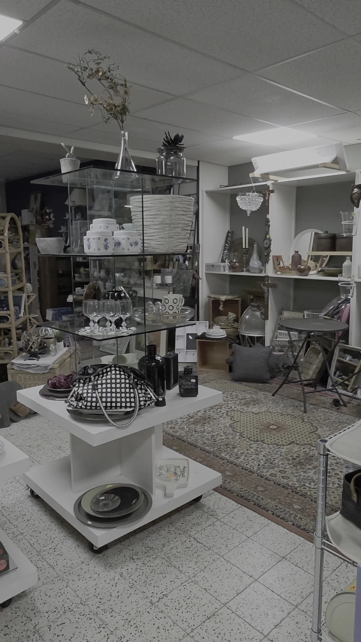 Winkelbinnenzicht met glazen vitrinekast met servies, decoratieve items op witte zuil, stellingen met huishoudspullen en meub