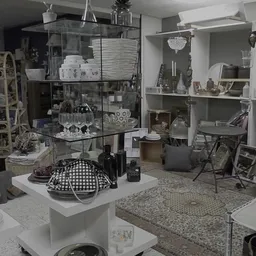 Winkelbinnenzicht met glazen vitrinekast met servies, decoratieve items op witte zuil, stellingen met huishoudspullen en meub