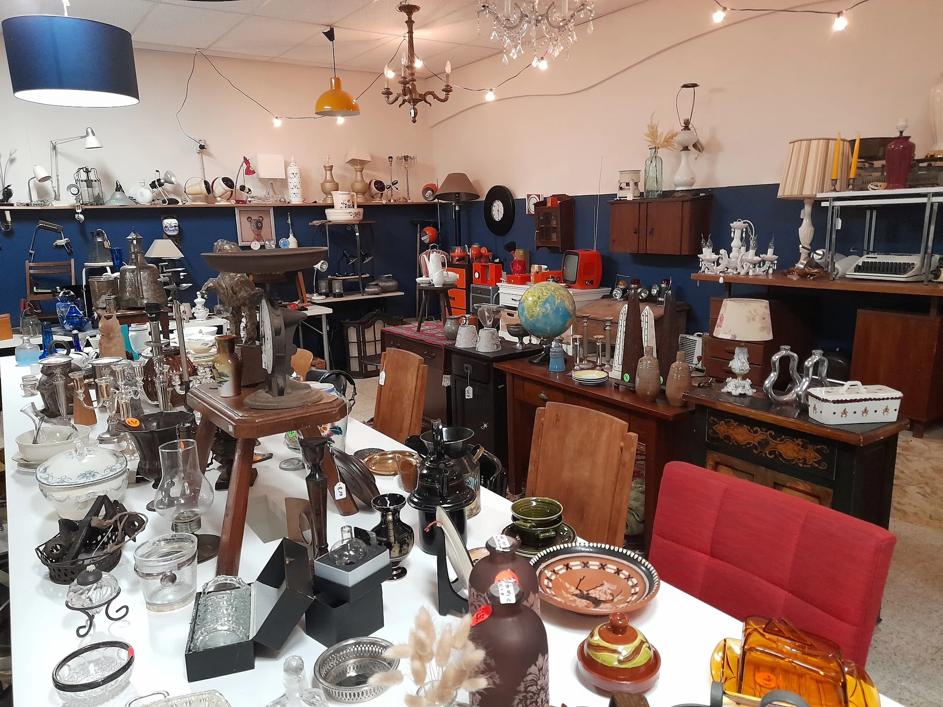 Ruim winkelbinnenruimte met diverse tweede-hands spullen, meubels, lampen en decoratieve objecten op tafels en stellingen.