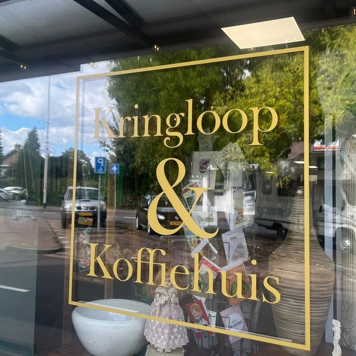 Gouden letters op winkelruit met tekst 'Kringloop & Koffiehuis', winkelinterieur en straat zichtbaar in spiegeling