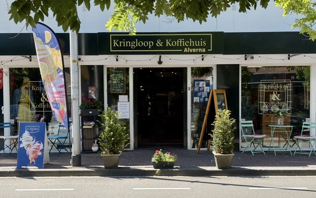 Winkelgevel met zwarte pui, witte omlijsting en gouden letters, potplanten aan ingang, terras met stoelen