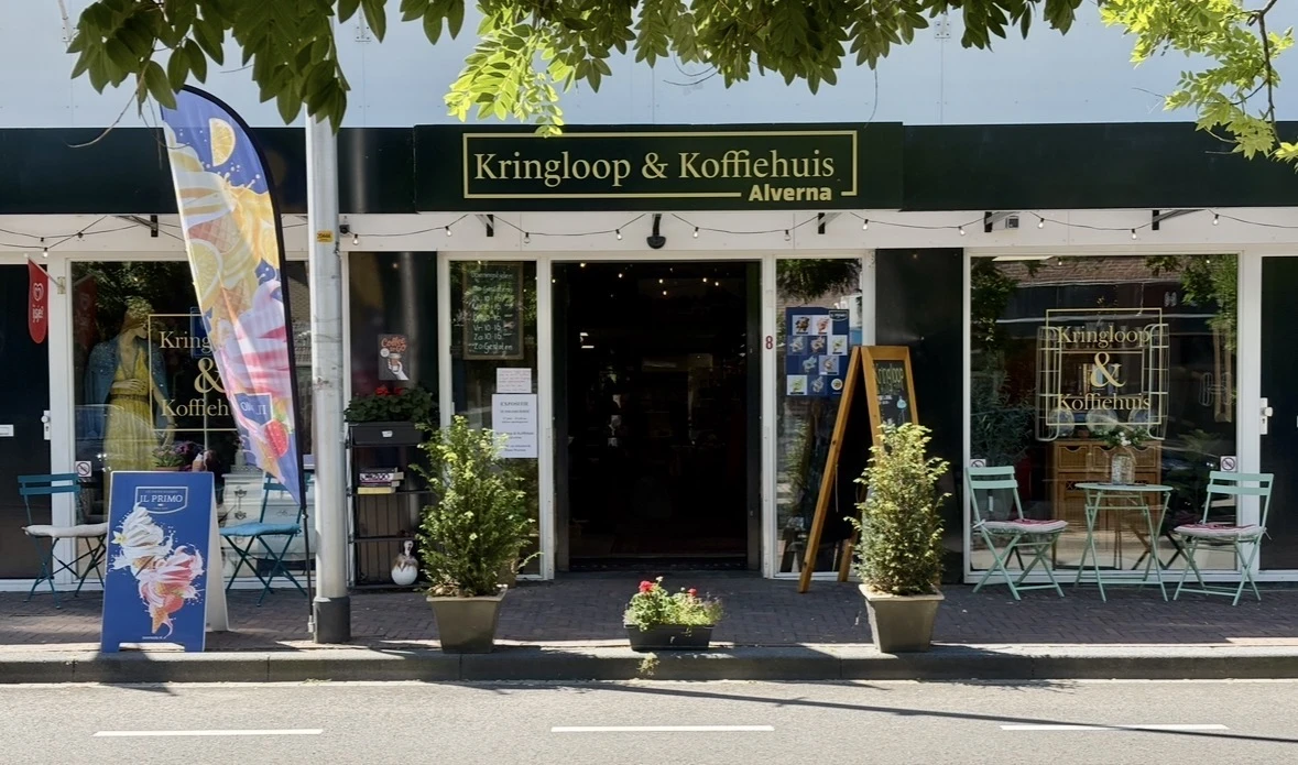 Winkelgevel met zwarte pui, witte omlijsting en gouden letters, potplanten aan ingang, terras met stoelen