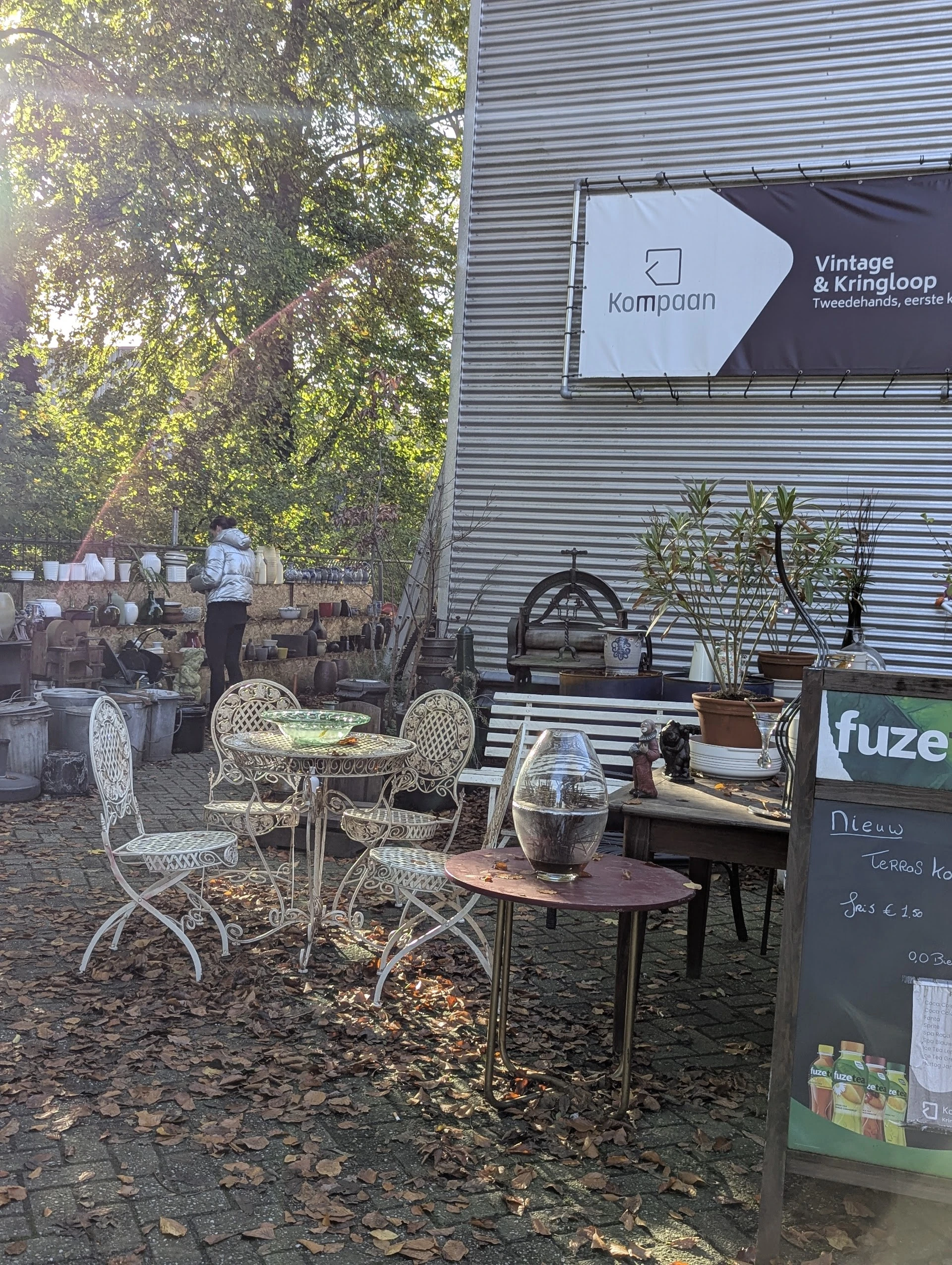Terras met witte stoelen, tafels en een bord met 'fuze' en menuopstelling buiten een kringloopwinkel
