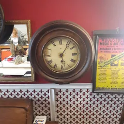 Vintage houten wandklok met Romeinse cijfers naast een gele Woodstock-festival poster, beide uitgestald in de winkel