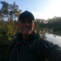 Man met pet en zwarte jas op de oevers van een rivier omgeven door bomen