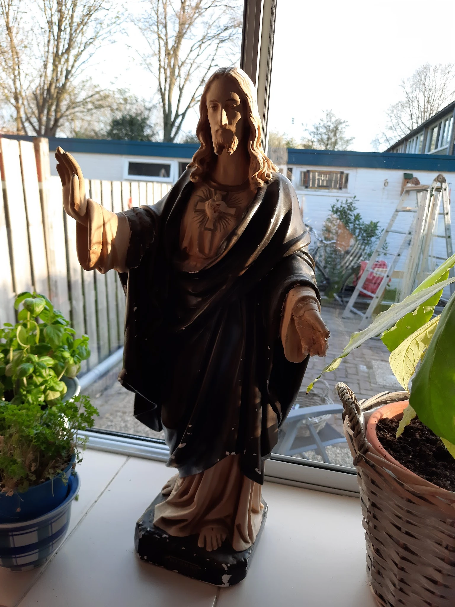 Bruinen houten beeldhouwwerk van een figuur in klassieke draperie, tegen raam met plantenkas op achtergrond