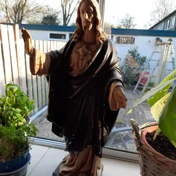 Bruinen houten beeldhouwwerk van een figuur in klassieke draperie, tegen raam met plantenkas op achtergrond