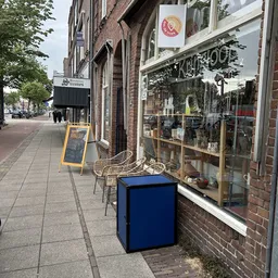 Straatgevel van kringloopwinkel met etalageruit, gele menukaart en blauw object op straat