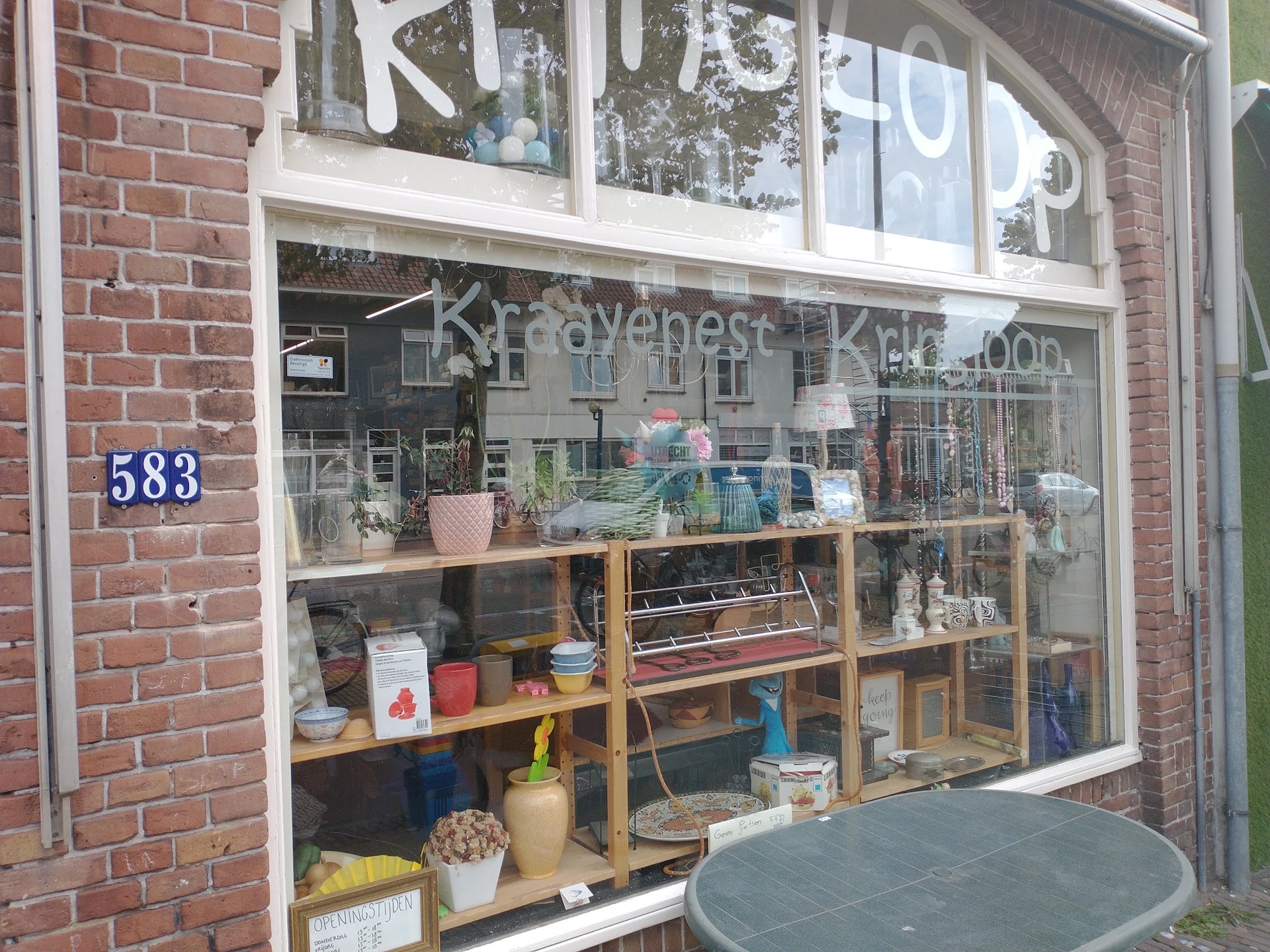 Winkelraam met vitrine van kringloopwinkel, witte kozijnen, nummerbord 583, diverse spullen zichtbaar