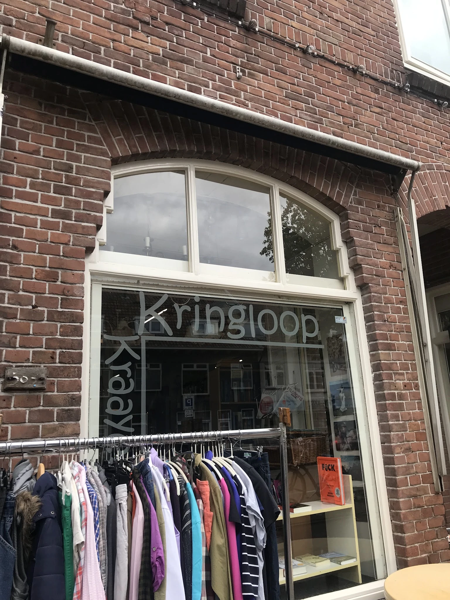 Gevel van kringloopwinkel met witte raamkozijnen en uithangbord, kledingrek met hangers zichtbaar in etalage