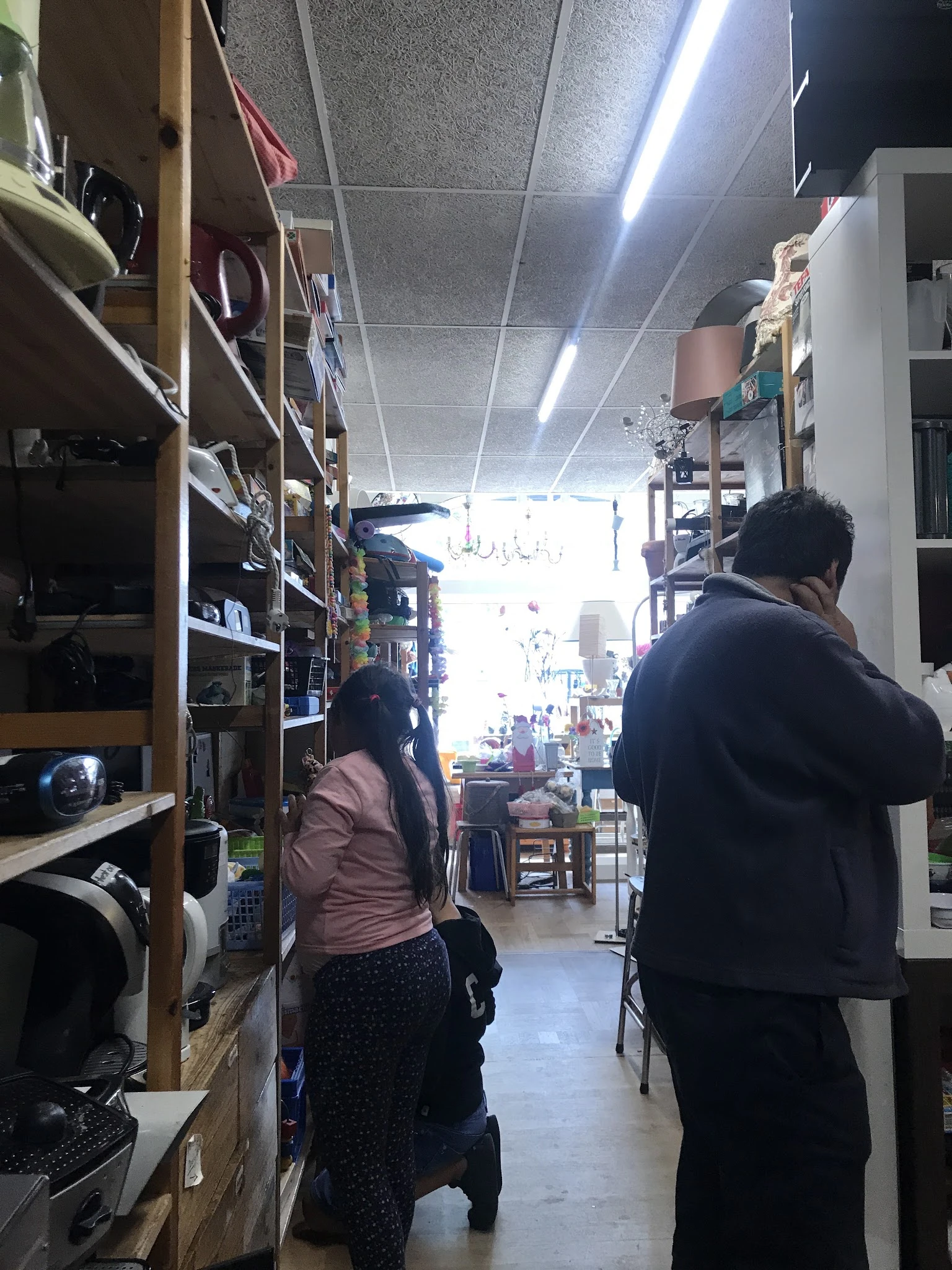 Winkelinterieur met houten stellingen vol spullen, twee bezoekers lopen door het middenpad naar de heldere voorkant