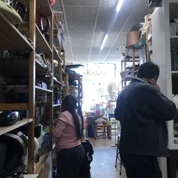 Winkelinterieur met houten stellingen vol spullen, twee bezoekers lopen door het middenpad naar de heldere voorkant
