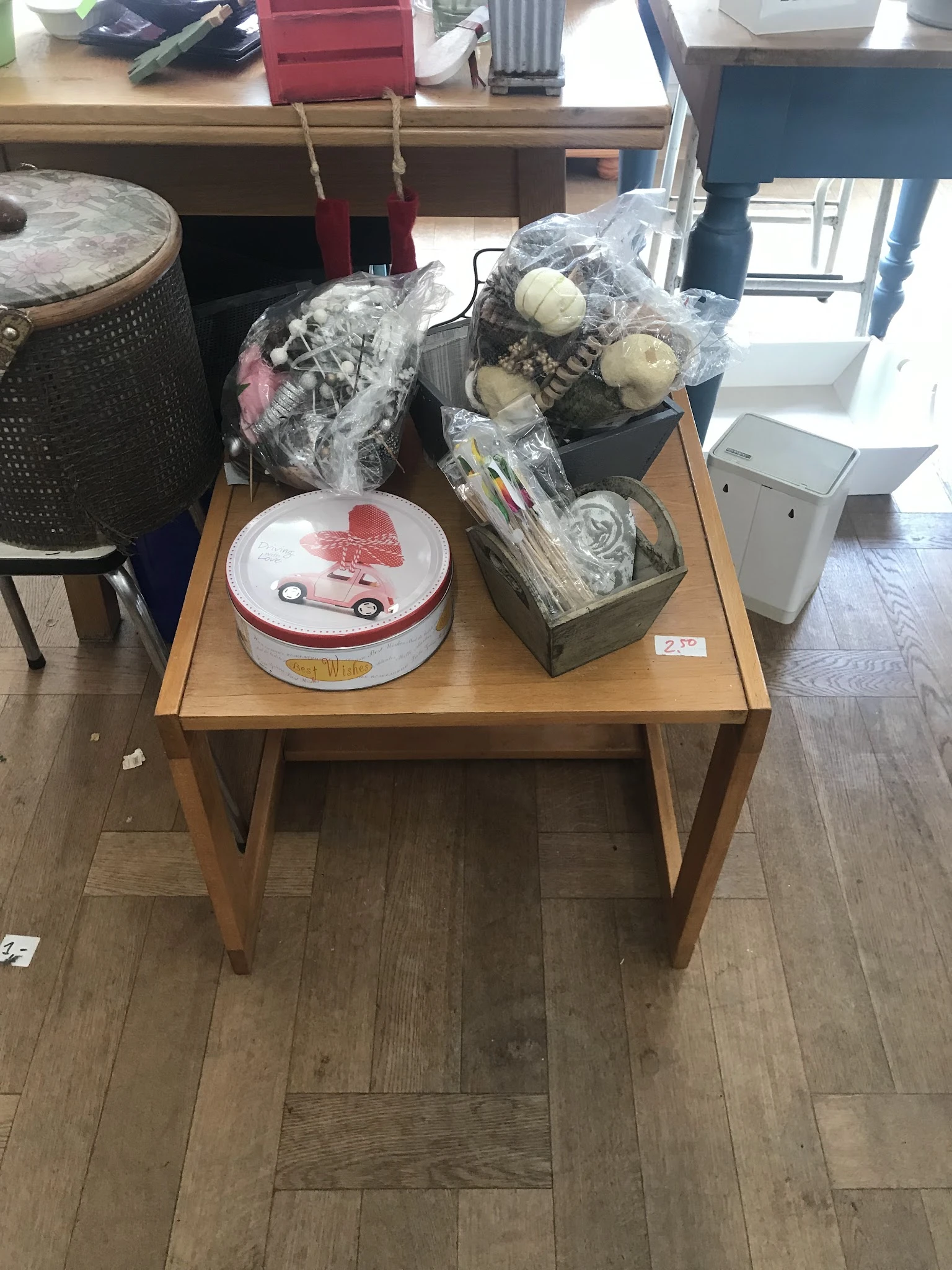 Houten tafel met diverse tweedehands artikelen: rode auto-deksel, plastic zakken met huishoudelijke spullen en een witte doos