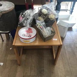 Houten tafel met diverse tweedehands artikelen: rode auto-deksel, plastic zakken met huishoudelijke spullen en een witte doos