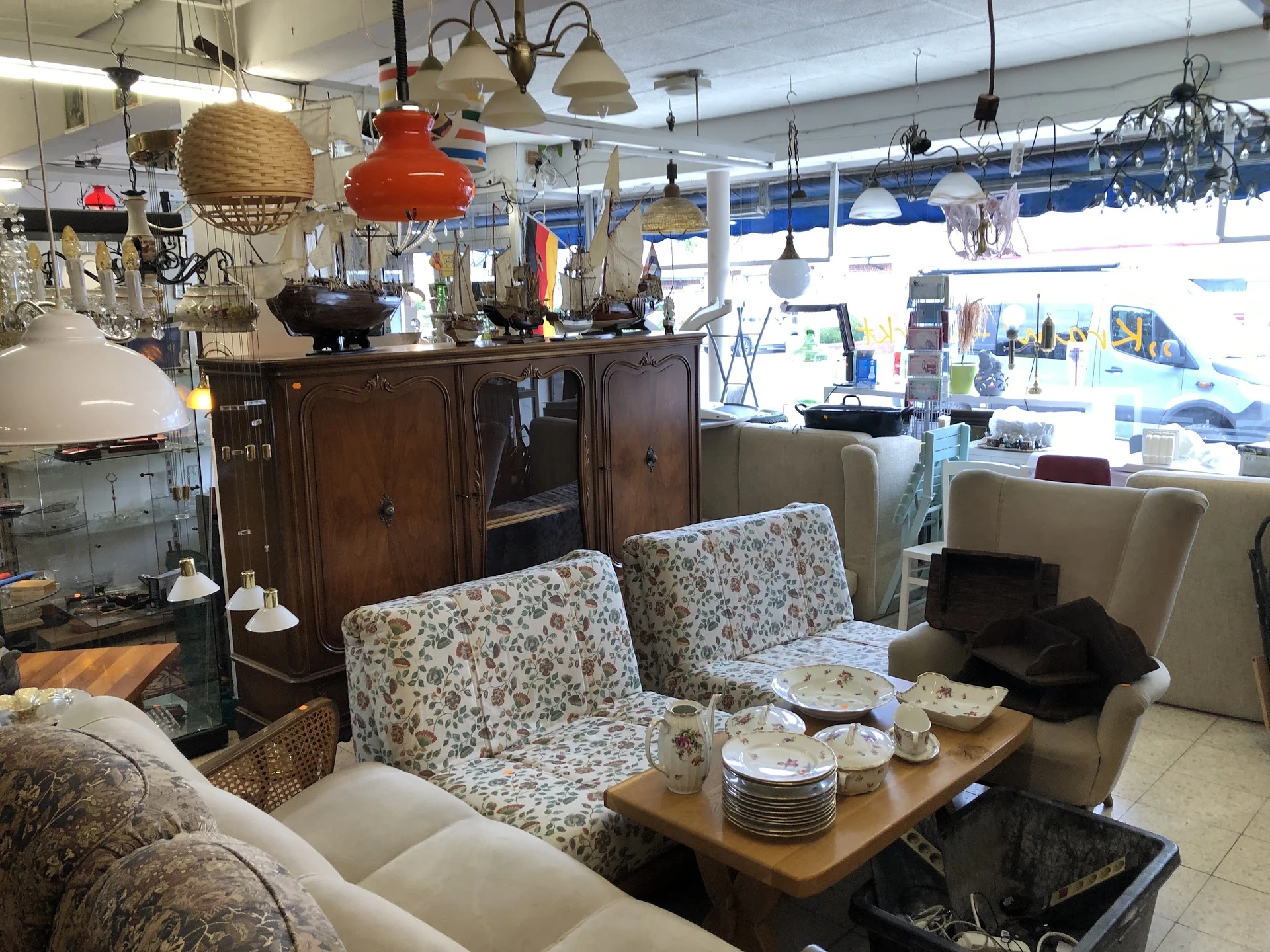 Winkelvloer met meubels: tweepersoonsbank, fauteuils, houten dressoir en hanglampen, achter grote ramen.