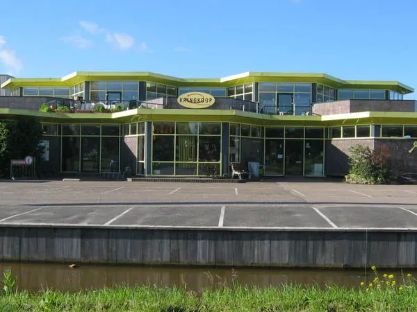 Moderne gevel van kringloopwinkel met groene afdaking, grote ramen en parkeerplaatsen ervoor