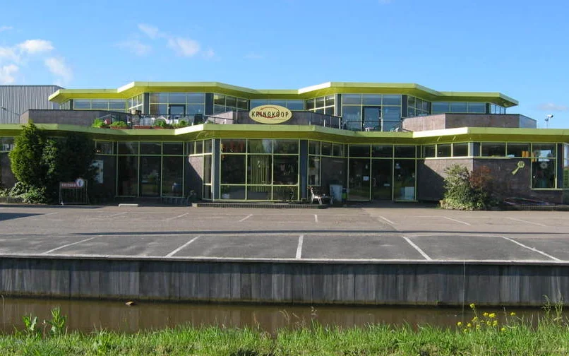 Moderne gevel van kringloopwinkel met groene afdaking, grote ramen en parkeerplaatsen ervoor