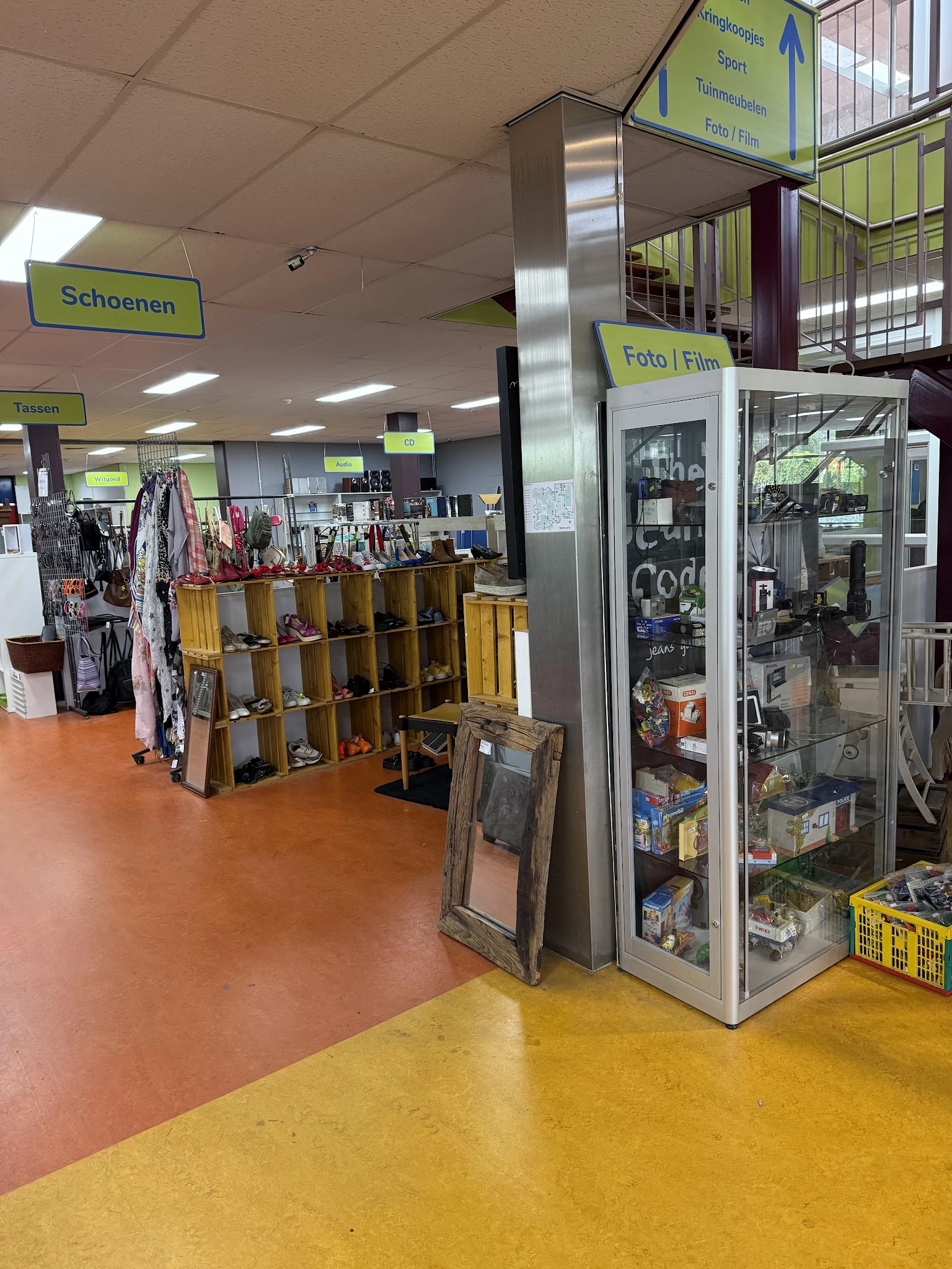 Winkelvloer met houten stellingen vol met kleding, schoenen en accessoires, fluorescerende gele borden en een glazen vitrinek