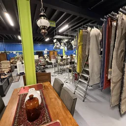 Winkelvloer met kleding op rekken, eettafel met servies en lampen in kringloopwinkel