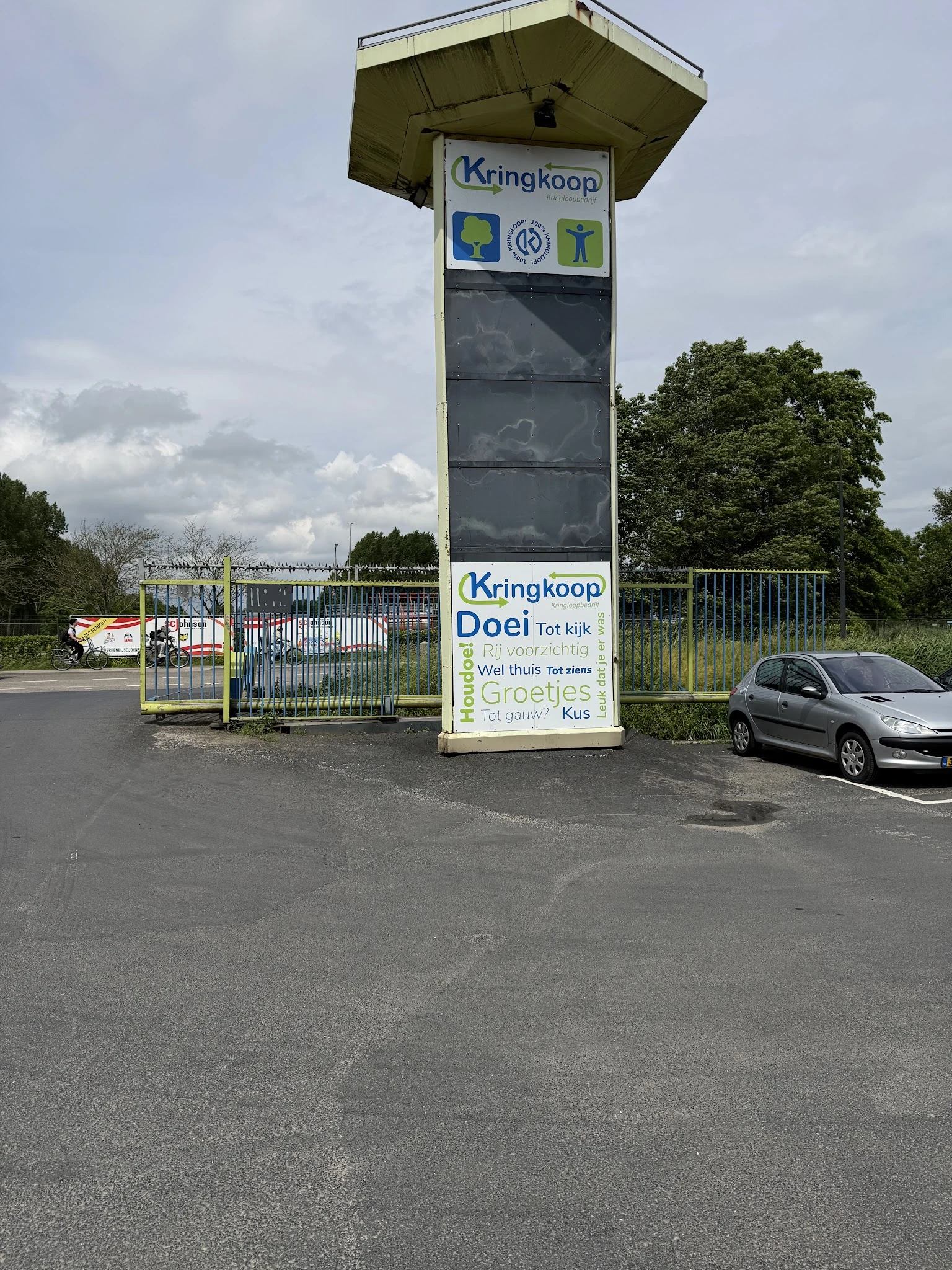 Ingang van kringloopwinkel met witte poort en groot bord met winzelnaam, auto geparkeerd, bomen op achtergrond