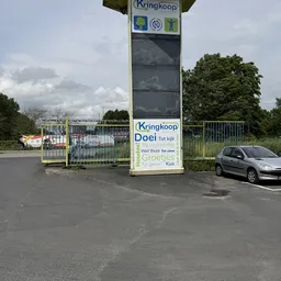 Ingang van kringloopwinkel met witte poort en groot bord met winzelnaam, auto geparkeerd, bomen op achtergrond