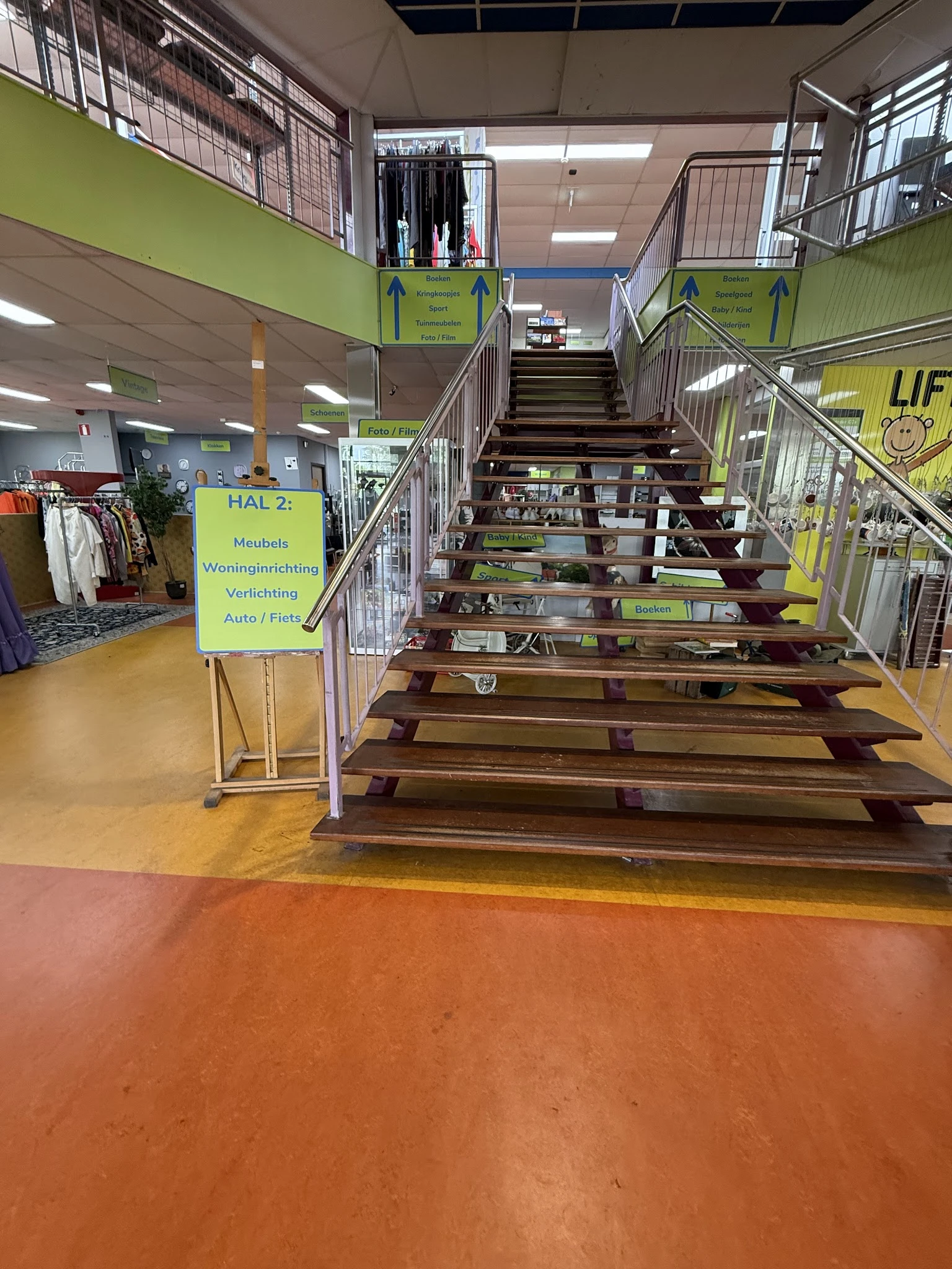 Trap naar bovenetage in kringloopwinkel met gele bewegwijzering en stellingen met spullen