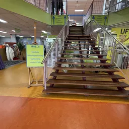 Trap naar bovenetage in kringloopwinkel met gele bewegwijzering en stellingen met spullen
