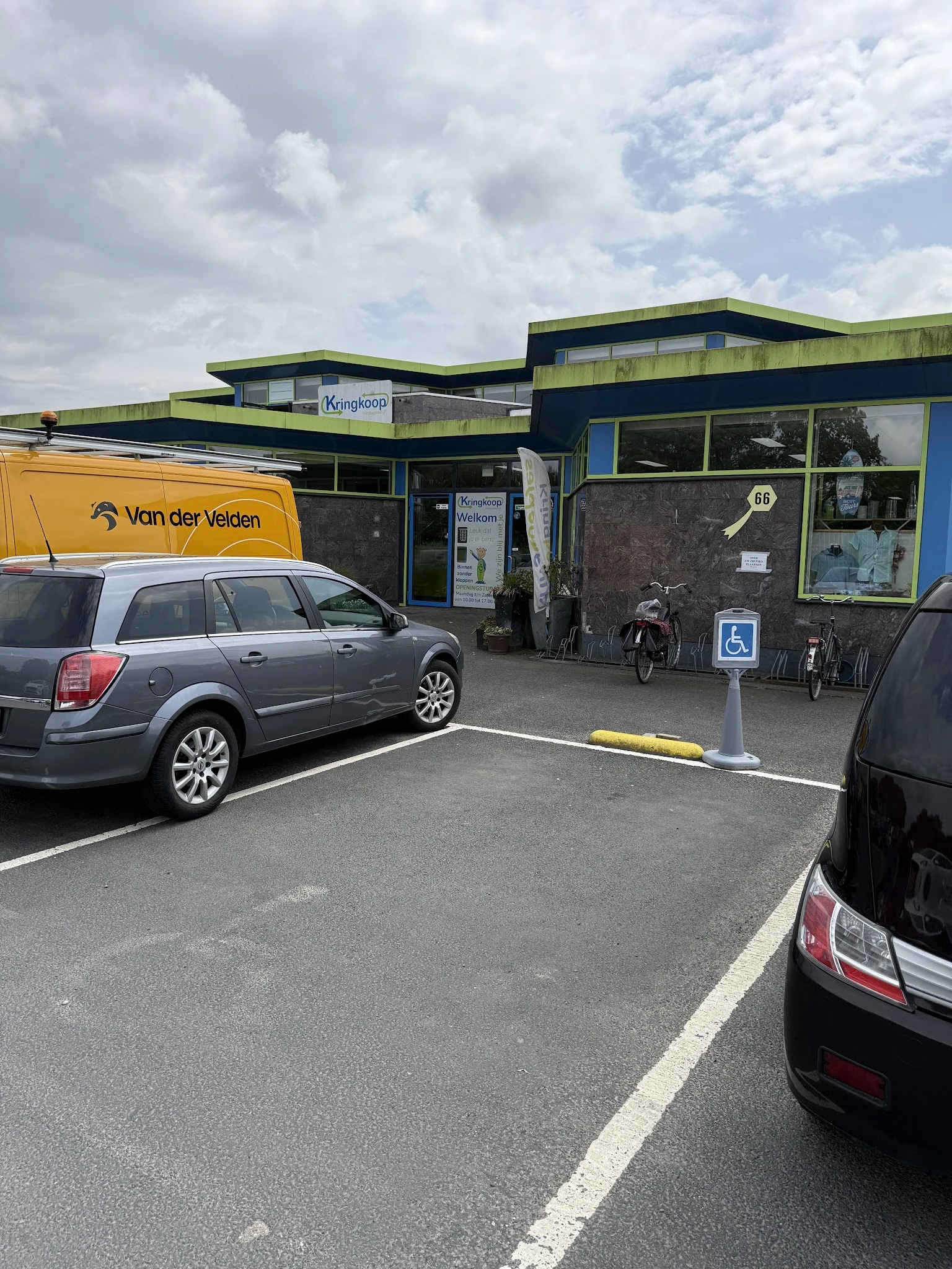 Voorkant van kringloopwinkel met groene en zwarte gevel, parkeerplaats met auto's en fietsen