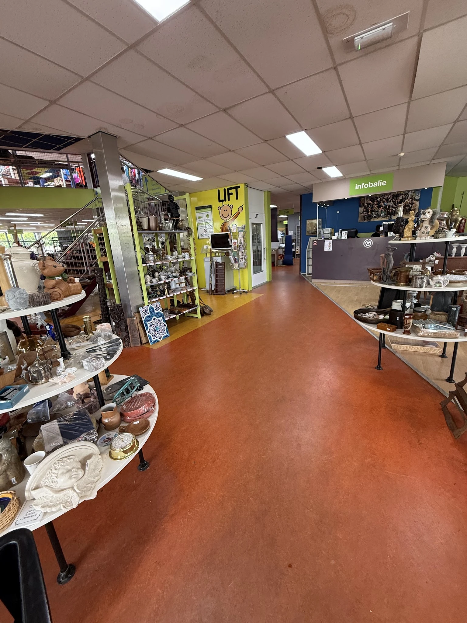 Winkelvloer van kringloopwinkel met diverse uitgestalde spullen, tafels met servies en decoratie, gele 'LIFT' deur en infobal