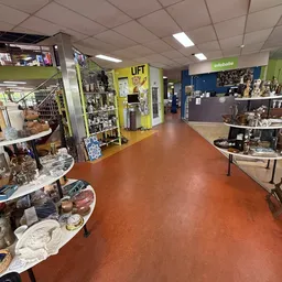 Winkelvloer van kringloopwinkel met diverse uitgestalde spullen, tafels met servies en decoratie, gele 'LIFT' deur en infobal